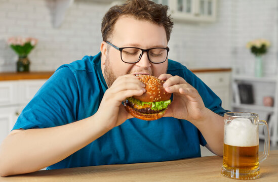 Overweight man holds burger and beer at home. Smiling plus-size adult earing unhealthy fast food and frothy mug, an indulgence at a wooden table in a kitchen. Cheat meal diet, obesity and nutrition.