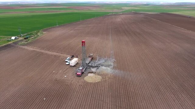 Water well drilling rig operating on farmland, aerial view of artesian well drilling process under clear sky. The machine penetrates deep into the ground for groundwater extraction and irrigation in a