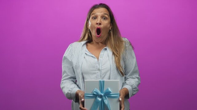 Woman holding a gift box tied with a blue ribbon, mouth open in surprise while presenting it to camera, studio; surprise celebration.