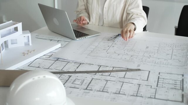 Architect working on new residential project designs, using laptop and reviewing blueprints with an architectural model and safety helmet on planning table