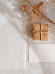 Fototapeta premium Clean minimal flat lay featuring a wrapped gift box, blank card, dried flowers, and parchment paper on a white marble surface for e-commerce presentation