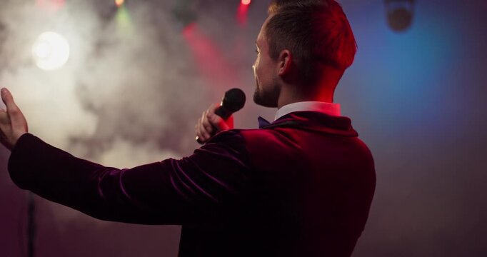 Host presenter showman with microphone on stage. Emcee and speaker engages crowd under lights and haze, ideal for concert promos, events, and nightlife ads. Clear show hosting and presentation.