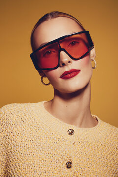 Stylish woman with slicked back hair wears oversized red sunglasses and gold hoop earrings. She poses confidently in a textured yellow sweater against a warm orange background.