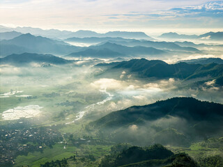 Obraz premium Panoramic aerial view of misty mountain range and rural village nestled in green valley at sunrise, highlighting serene natural beauty and peaceful morning atmosphere.