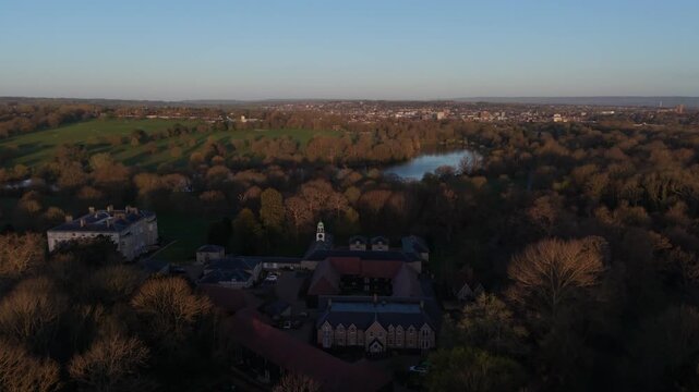 Mote Park in Maidstone with lake and historic buildings at sunrise.  Aerial view flying above the public park. Historic buildings and a clock tower in the sun. Maidstone town skyline at the distance. 