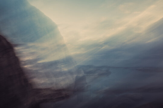 Dreamlike motion blurred coastal scene with soft pastel light, water, rocks and a ghosted human figure.