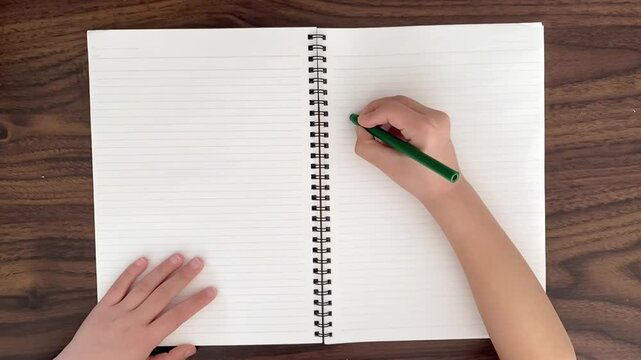 Child drawing a straight horizontal line in notebook with green pen on wooden desk, learning writing practice, slow motion