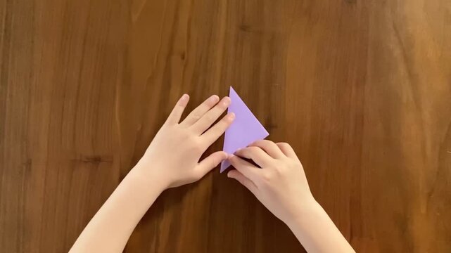 Child folding a small triangle origami into another triangle on wooden table, step by step paper folding tutorial, slow motion