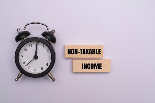 An alarm clock next to wooden blocks with text "NON-TAXABLE INCOME" on a white background. Concept for tax-free investments, retirement planning, and financial deadlines