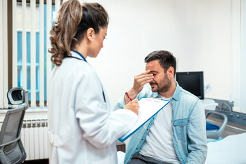 Obraz premium Male patient with headache talking to doctor in clinic. Professional clinician taking notes on clipboard during medical consultation. Healthcare and medicine concept, stress, migraine, and diagnosis.