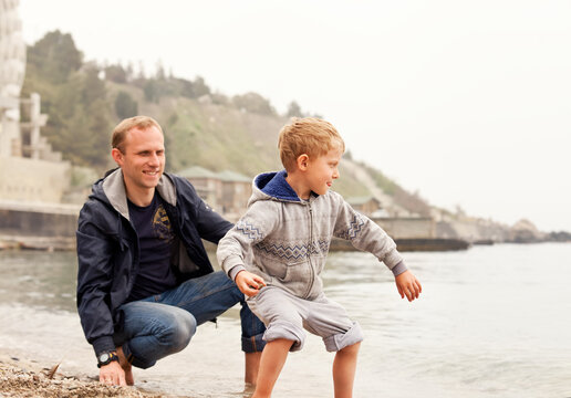 smiling father and young son playing together and skipping stones on water at pebble beach as concept of family fun, active lifestyle, and stone skimming during summer vacation at coastal resort