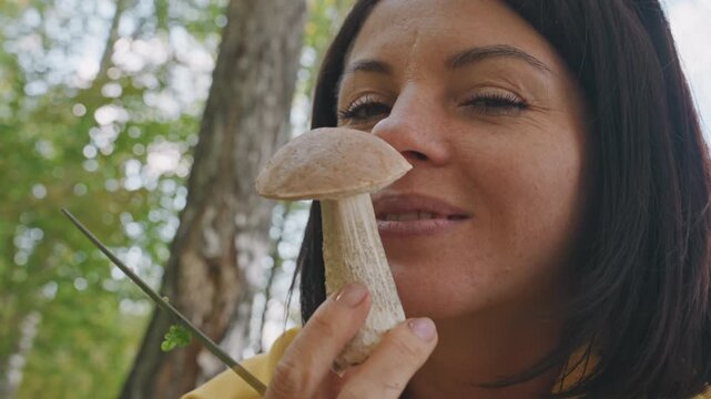 Woman inspecting mushroom during camping, smiling and smelling brown cap in sunlit forest hiker and amateur mycologist examines stem and texture with knife, careful inspection, quiet joyful moment