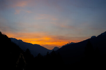 Obraz premium Spectacular sunset over a dark mountain range with silhouetted pine trees. Vibrant orange and yellow sky contrasting with dark alpine peaks. Mountain sunset background wallpaper 