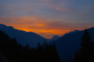 Obraz premium Spectacular sunset over a dark mountain range with silhouetted pine trees. Vibrant orange and yellow sky contrasting with dark alpine peaks. Mountain sunset background wallpaper 
