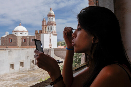 Young Latina woman applying makeup by window with church view in Mexico