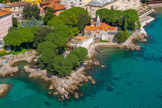 Aerial view of rocky coastline with small Church of St. James (Crkva sv. Jakov) and dense pine trees in Opatija, clear Adriatic water surrounding a scenic coastal walkway