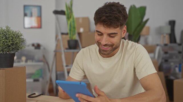 Man taps tablet screen with finger at wooden desk in building with moving boxes and potted plants; settling contentment.