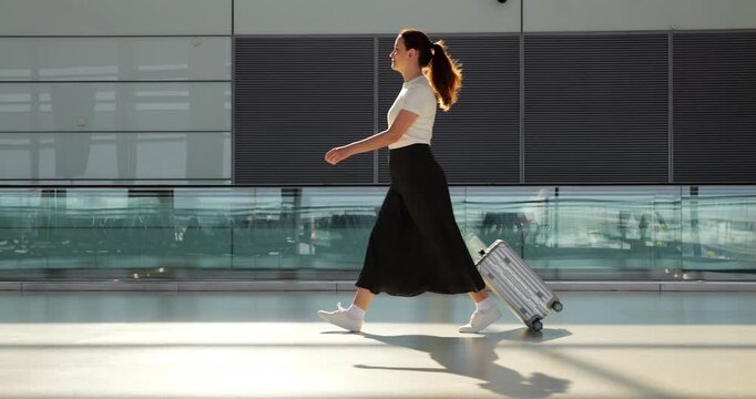 Elegant woman in long black skirt walking with her trolley case through airport terminal, side slow motion view. She confidently come alone to boarding gate, unhurried and steady.