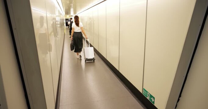 Rear view of a female passenger in slow motion, pulling a wheeled carry-on suitcase through a modern, minimalistic jet bridge with bright white walls and sleek, windowless interior design.