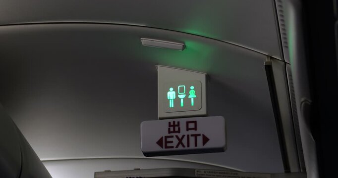 Low angle shot of glowing red lavatory occupancy sign on airplane ceiling console. illuminated indicator transitions from occupied light to green vacant light. Dim economy class cabin during flight.