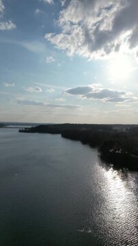 Vertical aerial drone view hovering over calm lake with sunlight reflection and clouds, peaceful nature background