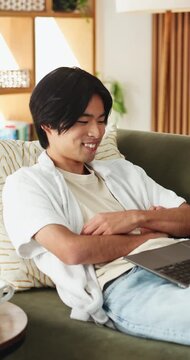 Streaming, laugh or Asian man in house with laptop, movie binging or funny video on online site. Happy, digital or person on sofa with tech, comedy show or series subscription on entertainment app.