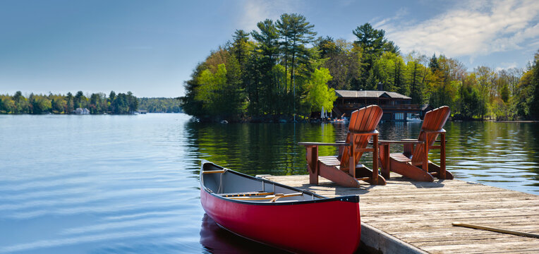 Two Adirondack chairs and a red canoe on a weathered Ontario dock face a calm Muskoka lake, boathouse and pine island;  the heart of Canadian cottage country in summer.