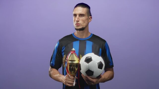 Man holding trophy and soccer ball in studio, wearing blue and black striped jersey and looking sideways; pride achievement teamwork.