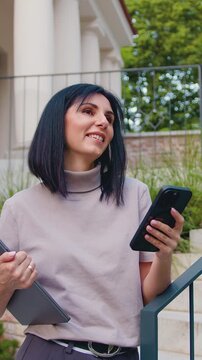 Pretty girl university student user holding cell phone and laptop using education tech apps looking at smartphone texting messages standing outside. Slow otion