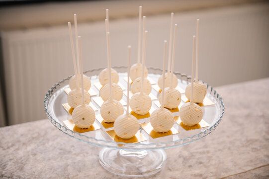 Elegant white chocolate cakesicles on sticks decorated with gold flakes. Professional dessert display on a glass stand for a wedding, anniversary, or sophisticated candy bar.