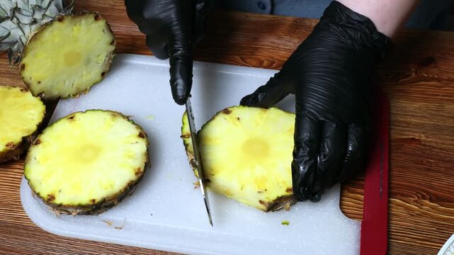 A chef wearing black gloves peels a pineapple with a knife on a cutting board.