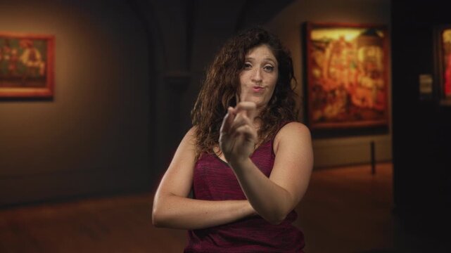 Woman points finger at viewer with arms crossed inside an art gallery building amid framed paintings and warm overhead lighting; skepticism assertiveness.