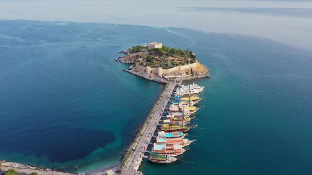 Aerial forward shot over Pigeon Island causeway in Kuşadası revealing the historic castle, colorful tour boats moored along the pier and turquoise Aegean waters