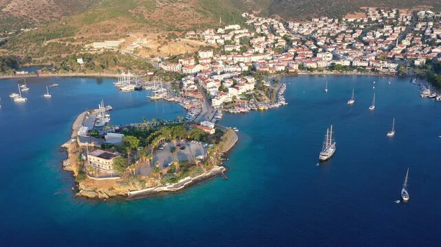 Panoramic aerial view of Datca harbor town with sailboats and a gulet anchored in a deep blue bay, marina and white-washed buildings climbing the hillside on a summer morning