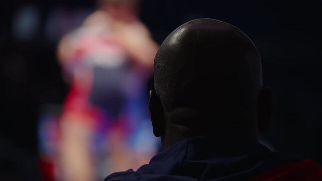 Silhouette of a focused coach watching two wrestlers grapple under dramatic arena lighting, capturing intensity, motion blur, and anticipation at the mat's edge during competition.