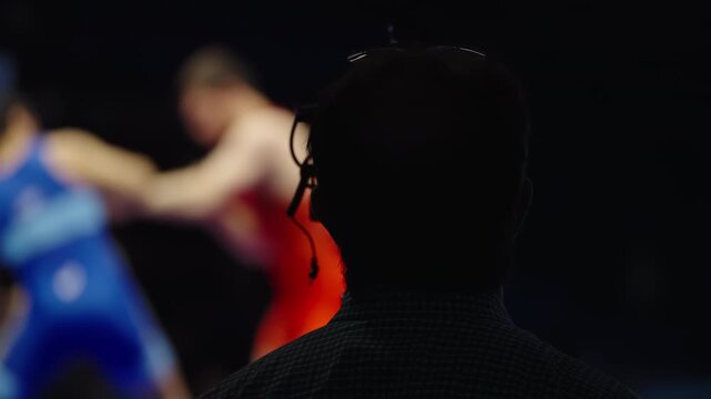 Silhouetted camera operator captures a wrestling match as two athletes in red and blue singlets grapple under arena spotlights, blending sports action with broadcast production