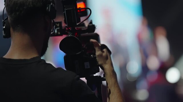 Professional videographer operating a shoulder-mounted camera with a headset during a live stage event, capturing performers under dramatic lighting with bokeh in the background.