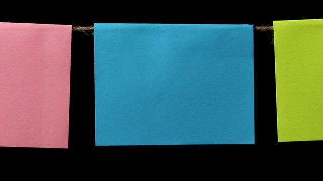 A square piece of blue paper and two other pieces of yellow and pink paper hang on a thin string against a black background. Space for text.