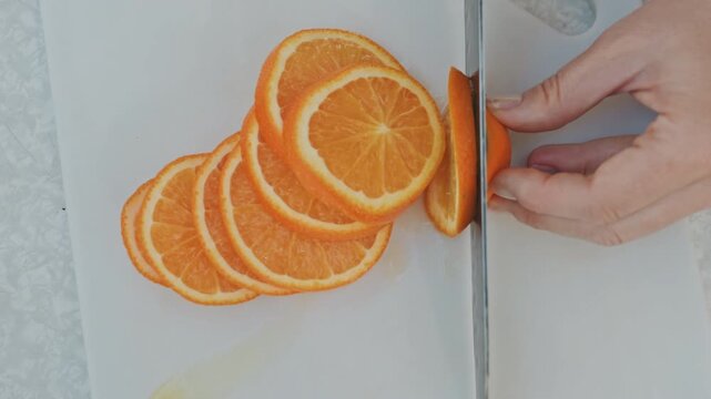 Overhead orange slices on white board, steady hands cutting thin rounds in row, white cutting board with droplets, sharp serrated blade, studio light, neat composition for recipe or styling, calm