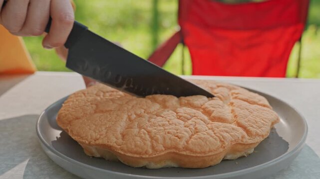 Outdoor camping table with bread being sliced by camper chef closeup serrated knife cutting soft cloud bread on plate red camp chair and green trees in background gentle morning light and simple