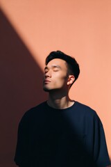 Fototapeta premium Male asian young adult sunlight profile peach background contemplative pose closed eyes dramatic shadow