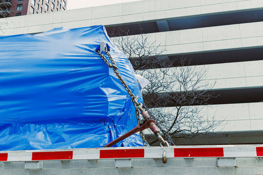 Large blue wrapped item is tied down on a truck near a parking structure in an urban area during daylight hours