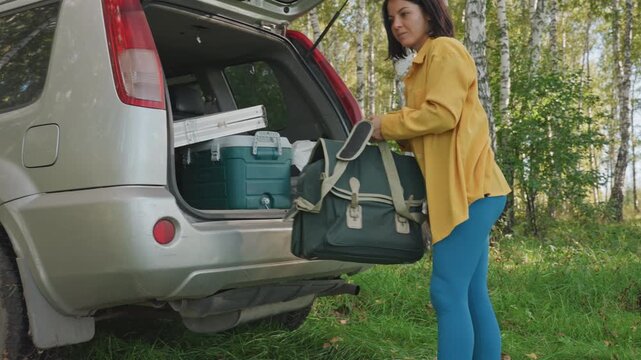 Woman loading cooler into car trunk, wearing yellow jacket and blue pants, carrying canvas bag next to suv in birch woodland, grassy campsite, closeup on bag on grass, midday sunlight, casual campsite