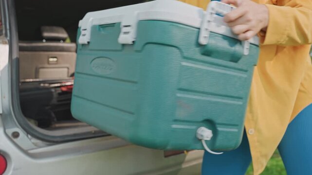 Person carefully handles outdoor cooler. Individual moves camping gear onto grassy area. Female outdoor enthusiast arranges camping equipment and refreshments with organized precision and care
