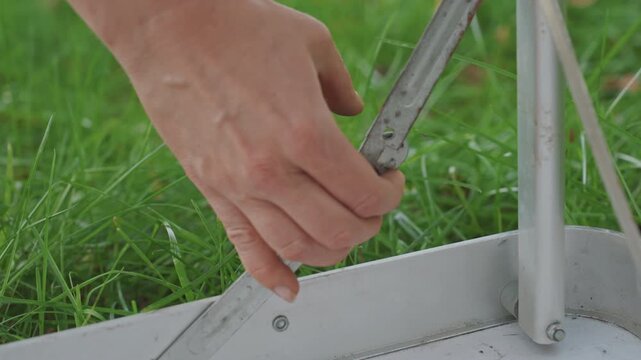 Closeup camping table hinge mechanics, hand adjusts metal bracket and rivet, detailed movement of pivot, signs of wear and rust, focus on maintenance and function, green grass background