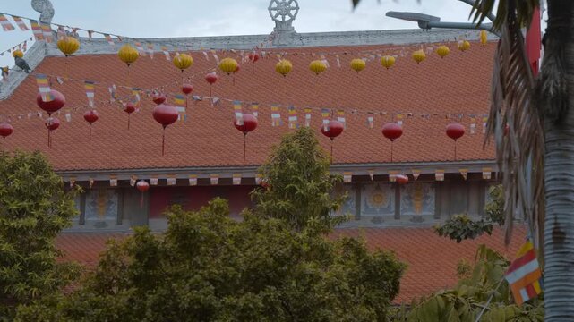 Garlands with Buddhist flags and colorful red and yellow Vietnamese lanterns flatter on the wind against the background of Vinh Nghiem Pagoda in Ho Chi Minh City, Vietnam with it's red tiled roof 