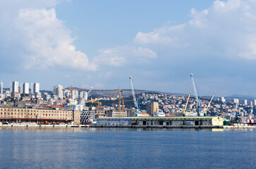 Obraz premium Panoramic view of an industrial port and coastal cityscape with cargo cranes and high-rise buildings under a cloudy sky.