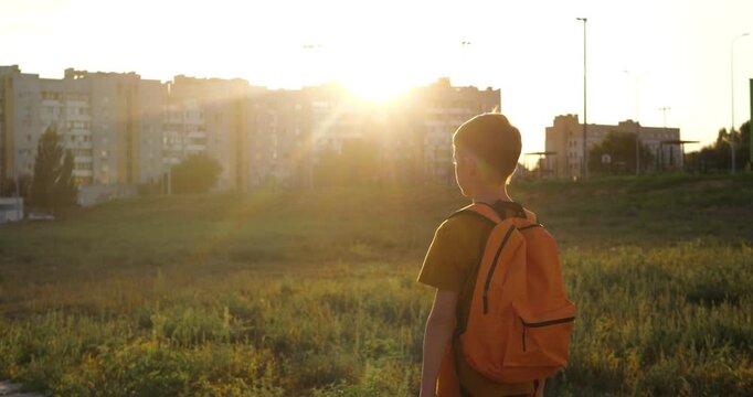 Boy with backpack walking outdoors in open urban field at sunset. Concept of childhood, freedom, and simple everyday life. Urban lifestyle child. child alone outdoors. boy sunset lifestyle