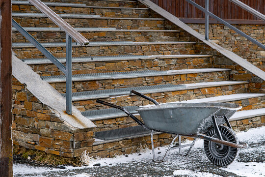Old metal wheelbarrow standing by stone stairs of a mountain shelter in winter, frosty day, frosted handrails and architectural details, maintenance work in cold conditions, harsh stone and ice textur