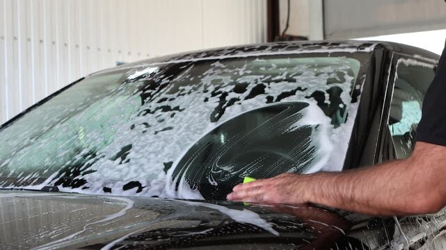 Male worker scrubbing soapy windshield with sponge at car wash, professional hand cleaning front glass with foam, auto detailing service